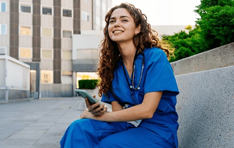 Enfermeira sorrindo, sentada ao ar livre, com o celular na mão