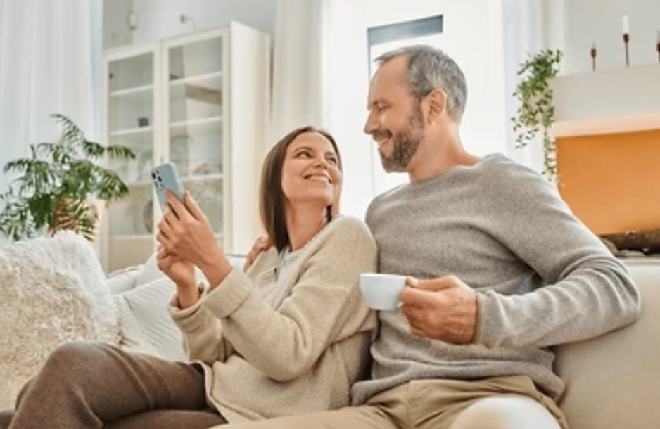 Casal no sofá de casa, sorrindo, a mulher mostrando uma mensagem no celular