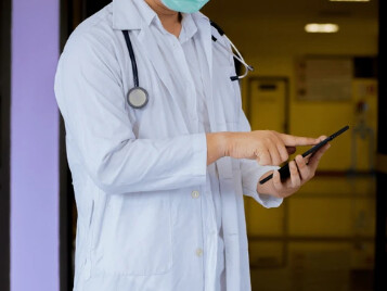 Profissional de jaleco e máscara consultando o celular, na porta do domicílio